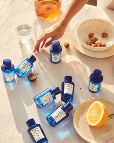 Eight blue Aqua di Parma bottles are strewn across a table with a white tablecloth next to a plates and bowls of fruit